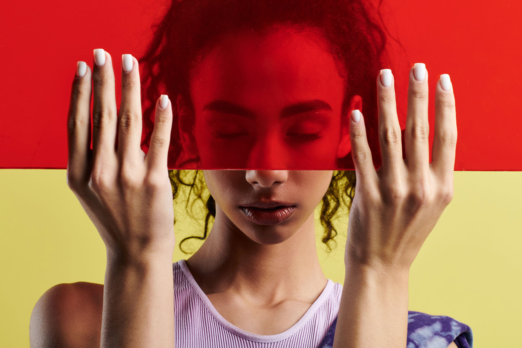 A young African American woman poses in a studio setting, holding a red panel in front of her face