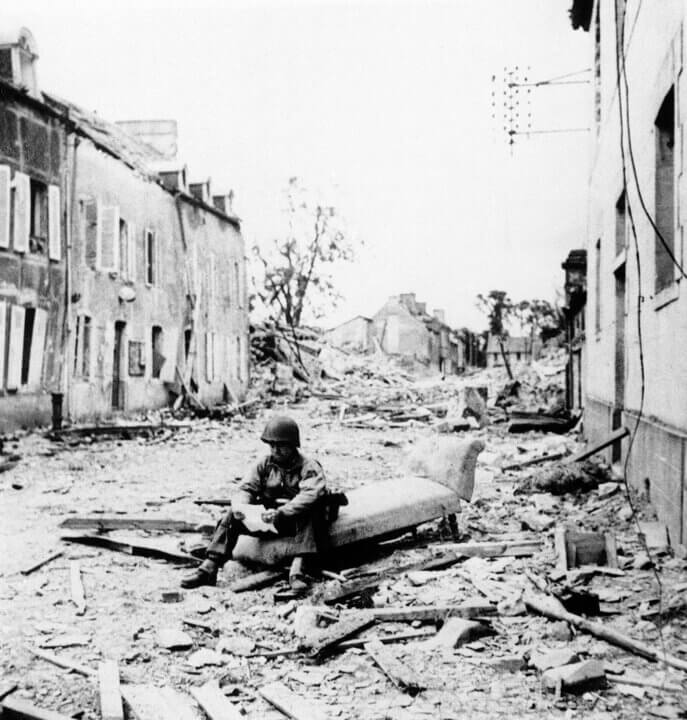 A soldier during The Second World War surrounded by bombed out building, sitting and reading a letter from his home in the States.