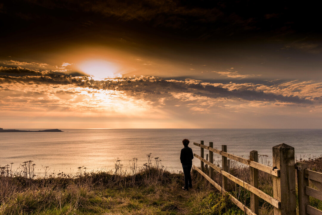A woman stands on cliffs looking at a sunset breaking over Newquay Bay in Cornwall.